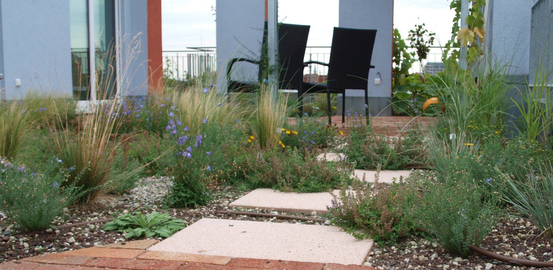 romantischer Sitzplatz auf Holzterrasse mit Blick über die Stadt, eingerahmt von Mauern mit Fenstern und Stauden