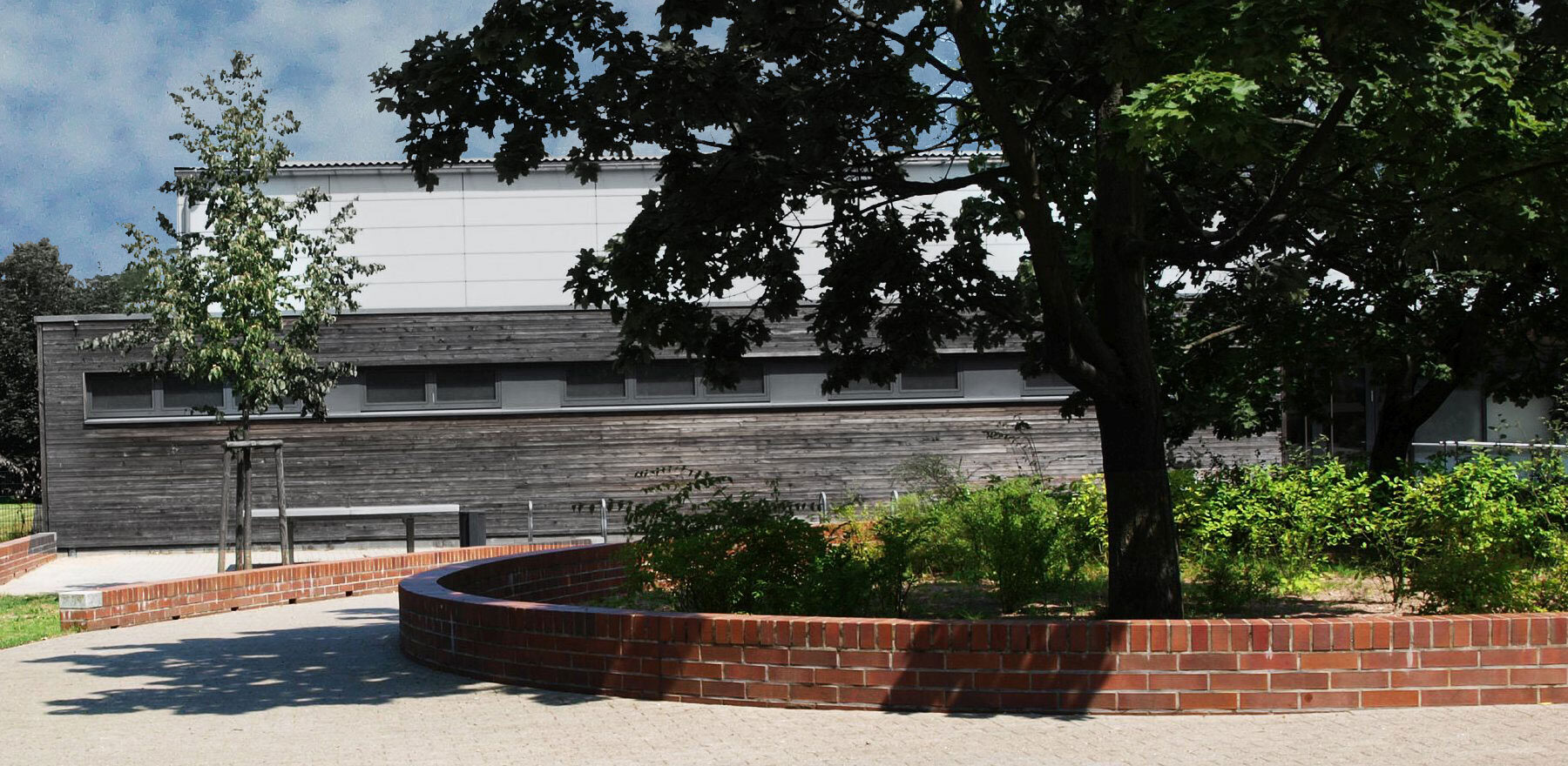 Frontalansicht Beeteinfassung aus rotem Klinker zum Sitzen um Baum und Sträucher vor Sporthalle